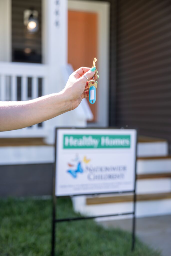 woman holding a key with her home in the background; she is standing in front of a Healthy Homes yard sign